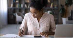 Serious young biracial woman sit at desk manage budget calculate on machine pay bills taxes online on laptop. Focused African American female count expenses expenditures on calculator.