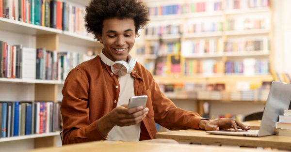 African American male student using cellphone and laptop sitting at desk in college library interior, texting while preparing for exams