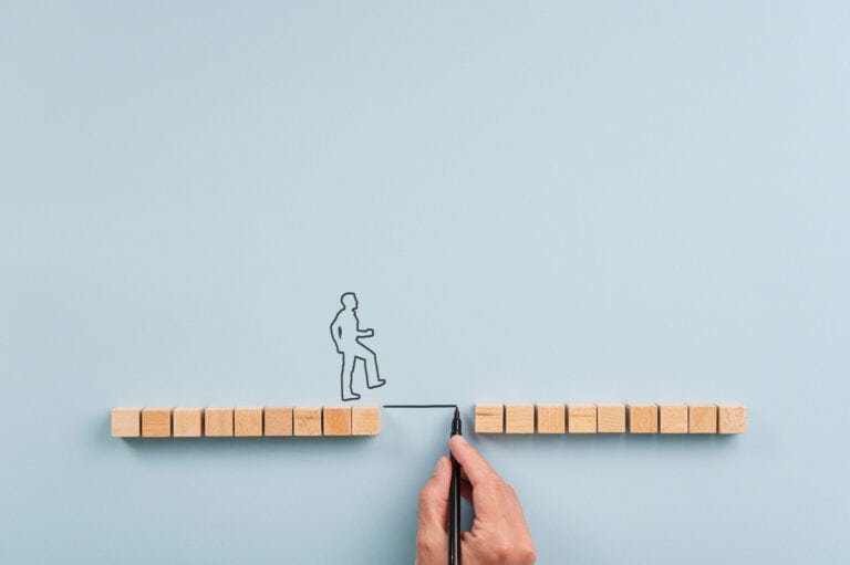 Male hand drawing a bridge between two rows of wooden blocks for a silhouetted man to walk across. Over blue background with plenty of copy space.